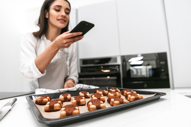Mulher jovem e atraente cozinhando saborosos biscoitos em uma bandeja enquanto está na cozinha, tirando uma foto
