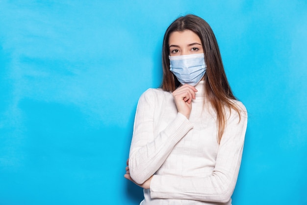 Mulher jovem e atraente de 20 anos com máscara facial protegida do vírus coronavírus covid-19 durante a quarentena pandêmica de mãos cruzadas, isolado em um fundo de cor azul brilhante.