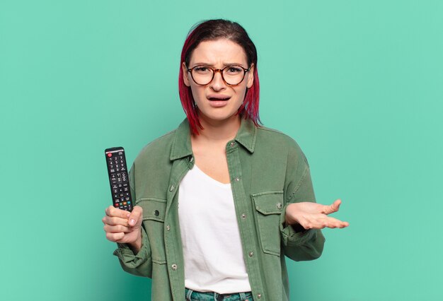 Mulher jovem e atraente de cabelo ruivo parecendo zangada, irritada e frustrada gritando wtf ou o que há de errado com você e segurando um controle remoto de TV