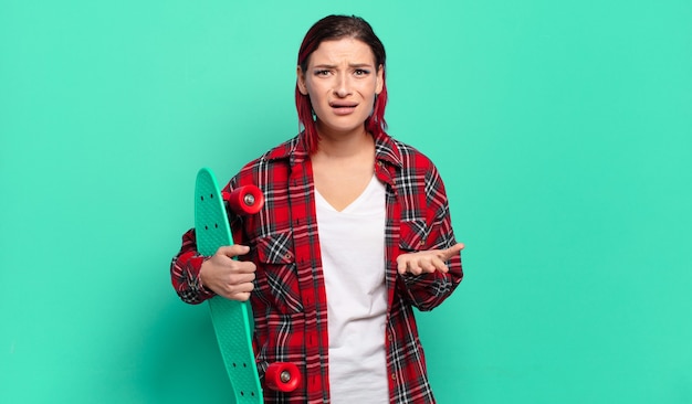 Mulher jovem e atraente de cabelo ruivo parecendo zangada, irritada e frustrada gritando wtf ou o que há de errado com você e segurando uma prancha de skate