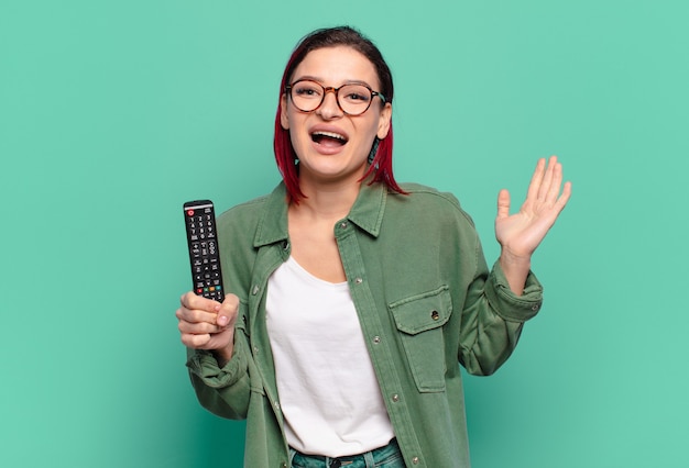 Mulher jovem e atraente de cabelo ruivo se sentindo feliz, animada, surpresa ou chocada, sorrindo e atônita com algo inacreditável e segurando um controle remoto de TV