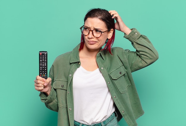Mulher jovem e atraente de cabelo ruivo se sentindo perplexa e confusa, coçando a cabeça e olhando para o lado segurando um controle remoto de TV