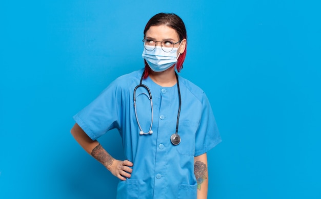 Mulher jovem e atraente de cabelo ruivo sorrindo alegremente e sonhando acordado ou duvidando, olhando para o lado. conceito de enfermeira de hospital