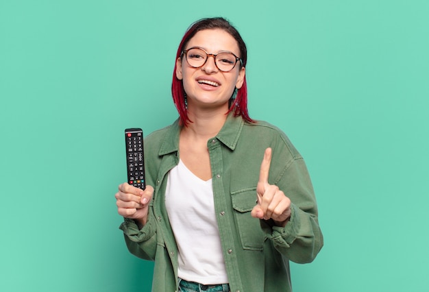 Mulher jovem e atraente de cabelo ruivo sorrindo com orgulho e confiança fazendo a pose número um triunfantemente, sentindo-se uma líder e segurando um controle remoto de TV