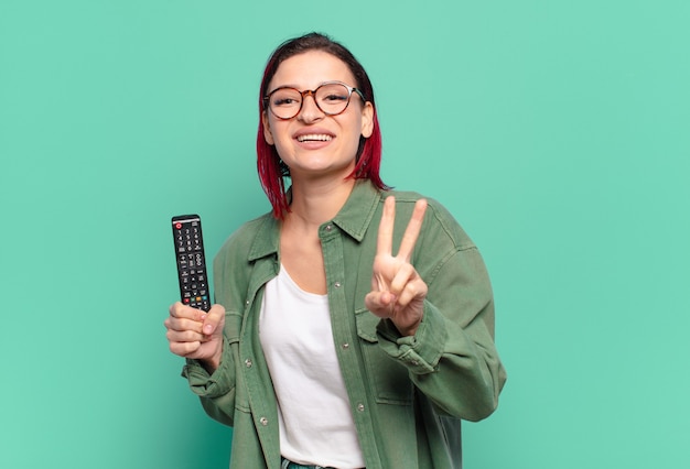 Mulher jovem e atraente de cabelo ruivo sorrindo e parecendo feliz, despreocupada e positiva, gesticulando para a vitória ou paz com uma das mãos e segurando um controle remoto da TV