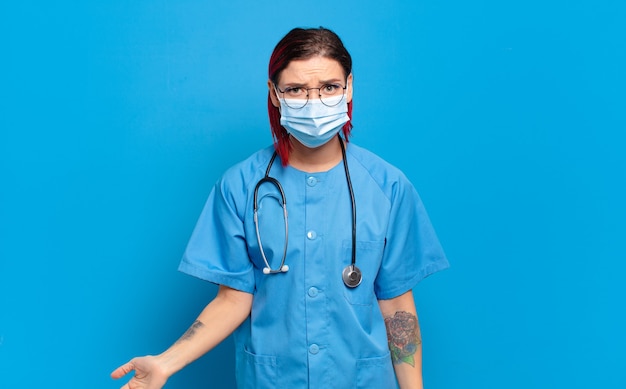 Mulher jovem e atraente de cabelo vermelho parecendo perplexa e confusa, mordendo o lábio com um gesto nervoso, sem saber a resposta para o problema. conceito de enfermeira de hospital