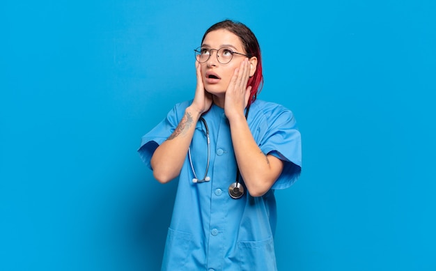 Mulher jovem e atraente de cabelo vermelho se sentindo feliz, animada e surpresa, olhando para o lado com as duas mãos no rosto. conceito de enfermeira de hospital