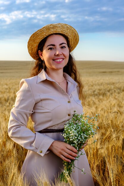 Mulher jovem e atraente de cabelos compridos com chapéu de palha sorrindo e segura um buquê de flores silvestres no campo de trigo ao nascer do sol.