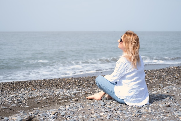 Mulher jovem e atraente de camisa branca e jeans azul com óculos de sol sentada na praia