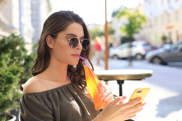 Mulher jovem e atraente em óculos de sol segura uma garrafa de suco, usando smartphone, ao ar livre.