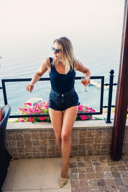 Mulher jovem e atraente em um top preto e shorts em um terraço com vista para o mar e bebe champanhe