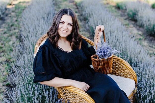 Mulher jovem e atraente em vestido preto, sentado na cadeira rodeada por campo de lavanda e segurando cesta com flores