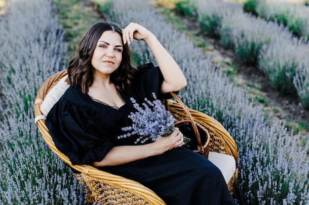 Mulher jovem e atraente em vestido preto, sentado na cadeira rodeada por campo de lavanda e segurando cesta com flores