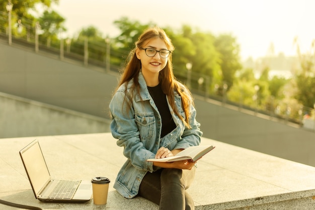 Mulher jovem e atraente estudante está lendo um livro enquanto está sentado na escada na cidade. O conceito de ensino à distância. Estudante moderno