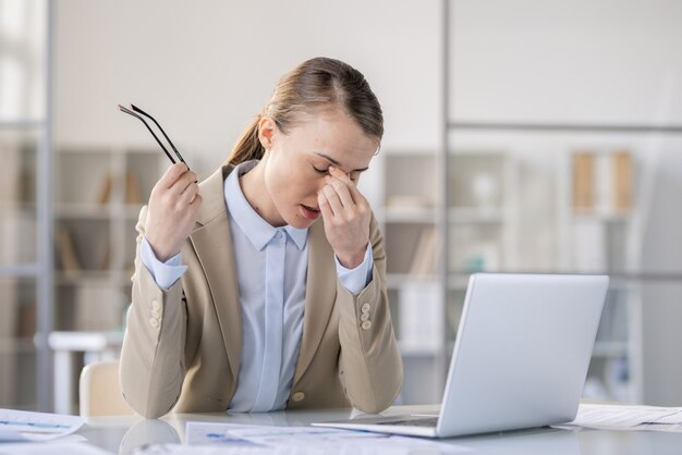 Mulher jovem e atraente exausta com uma jaqueta cansada do trabalho com o computador sentado à mesa e esfregando a ponte do nariz enquanto tirava os óculos, conceito de fadiga ocular
