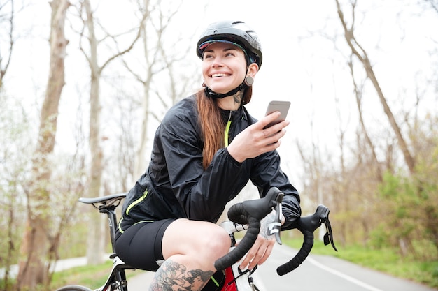 Mulher jovem e atraente feliz em capacete de ciclismo em bicicleta usando smartphone