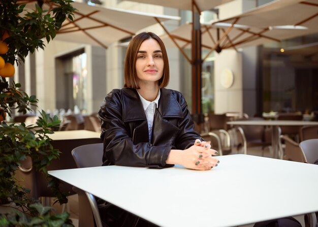 Mulher jovem e atraente linda morena confiante com corte de cabelo curto em uma jaqueta de couro preta ecológica, sentada à mesa no terraço de um café aconchegante, olhando para a câmera com confiança