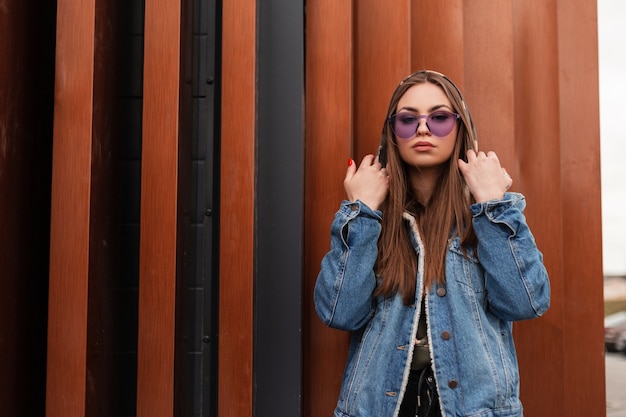 Mulher jovem e atraente moderna no elegante casaco jeans azul com capuz militar com óculos roxos da moda se passando perto de uma parede de metal vintage ao ar livre. Menina da moda hippie na cidade. Estilo de rua.