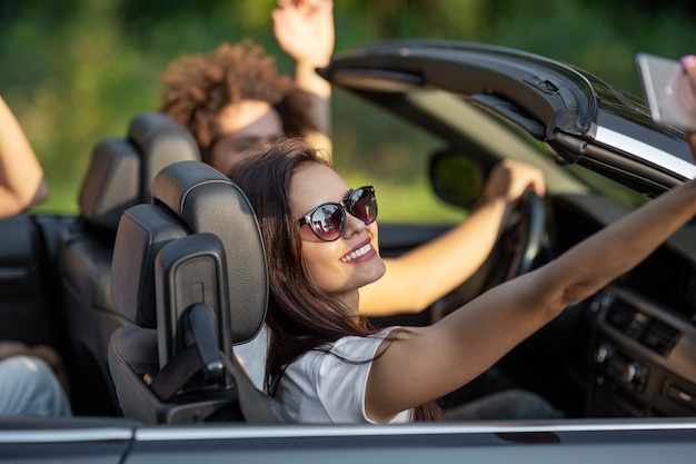 Mulher jovem e atraente morena de óculos escuros se senta com os amigos em um sorriso preto cabriolet e fazer uma selfie em um dia ensolarado. .