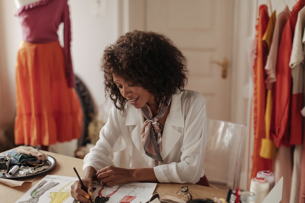 Mulher jovem e atraente, morena, encaracolada, de pele escura, com blusa branca da moda projeta coleção de moda e se senta em um escritório aconchegante