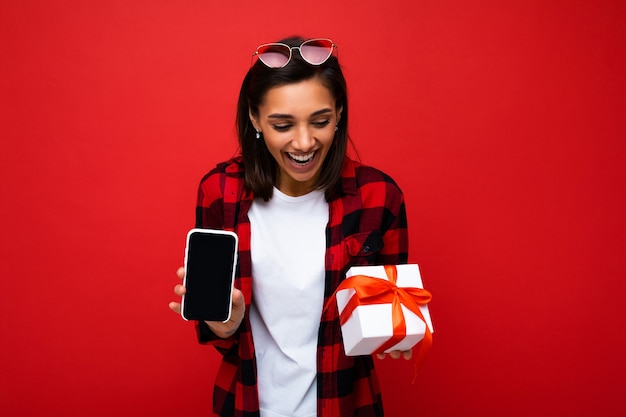 Mulher jovem e atraente morena feliz isolada sobre uma parede de fundo vermelho usando uma camiseta casual branca