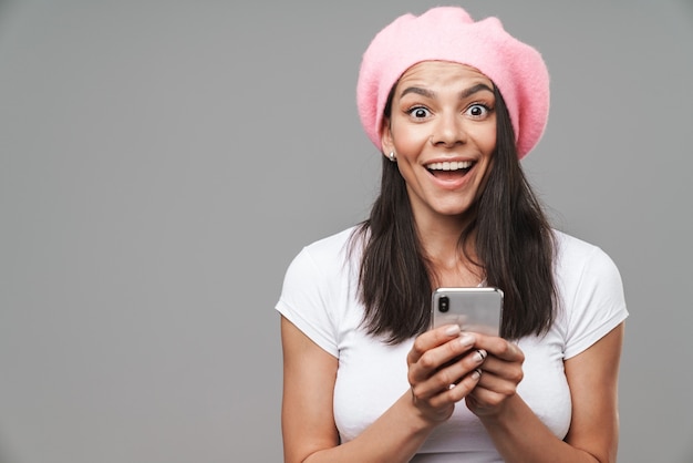 Mulher jovem e atraente morena feliz usando uma boina em pé, isolada na parede cinza, segurando um telefone celular, comemorando o sucesso