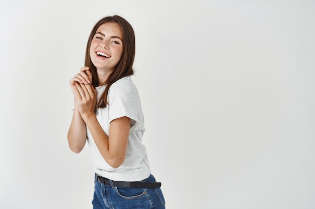 Mulher jovem e atraente morena rindo e olhando feliz na frente, posando contra uma parede branca.