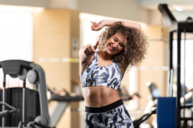 Mulher jovem e atraente mostrando os polegares para cima enquanto está forte no ginásio e flexionando os músculos Belo modelo de aptidão atlética posando após exercícios