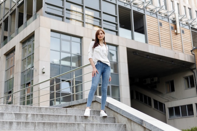 Mulher jovem e atraente na cidade. Mulher de negócios em pé perto do centro de negócios. Mulher de negócios cansada com uma camisa branca e calça jeans sentada relaxando na rua perto do prédio de vidro do centro da cidade