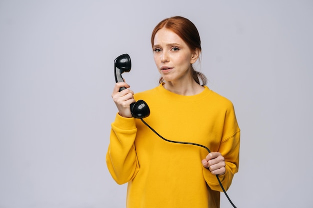 Mulher jovem e atraente no elegante suéter amarelo falando no telefone retrô e olhando para a câmera