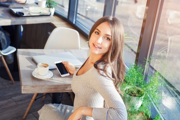 Mulher jovem e atraente no vestido no café usa celular e laptop