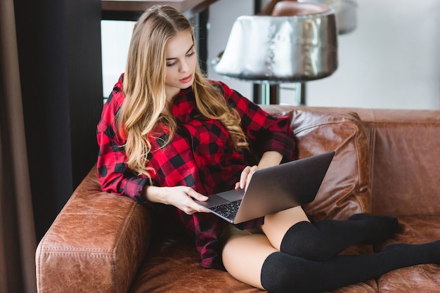 Mulher jovem e atraente pensativa com camisa quadriculada e meias pretas relaxando no sofá de couro e usando o laptop