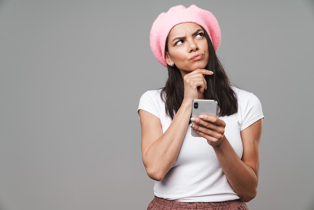 Foto mulher jovem e atraente, pensativa, morena, usando uma boina em pé, isolada na parede cinza, segurando um telefone celular