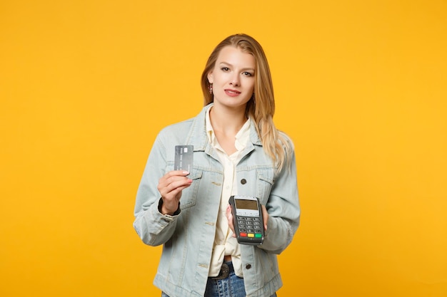 Mulher jovem e atraente segurando o terminal de pagamento bancário moderno sem fio para processar e adquirir pagamentos com cartão de crédito isolados em fundo amarelo laranja. Conceito de estilo de vida de pessoas. Mock up espaço de cópia.