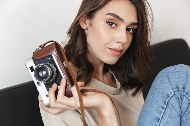 Mulher jovem e atraente segurando uma câmera fotográfica retrô e sentada no sofá do apartamento