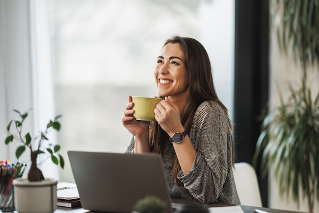 Mulher jovem e atraente sentada e segurando uma xícara de chá enquanto usa seu laptop em casa.