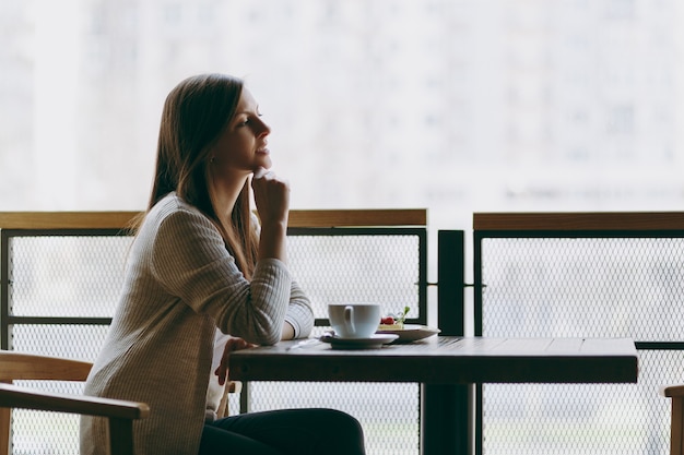 Mulher jovem e atraente sentada sozinha perto da grande janela na cafeteria na mesa com uma xícara de cappuccino, bolo, relaxante no restaurante durante o tempo livre. Jovem fêmea a descansar no café. Conceito de estilo de vida.