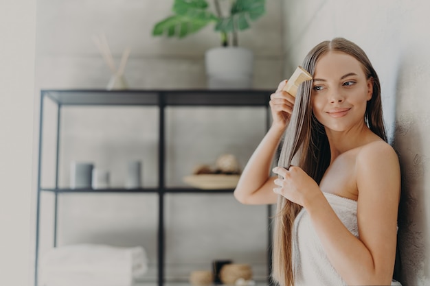 Mulher jovem e atraente sorri e aprecia sua beleza, penteia o cabelo longo e reto após a aplicação da máscara, fica enrolada em toalha de banho no banheiro aconchegante, tem pele macia.