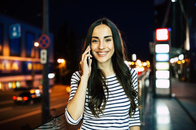 Mulher jovem e atraente sorridente fala ao telefone no fundo da cidade à noite