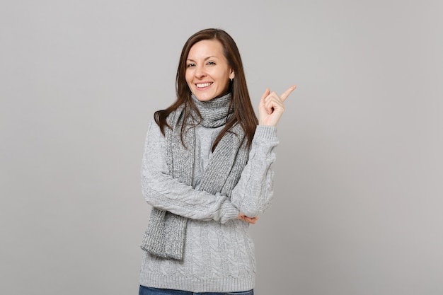 Mulher jovem e atraente sorridente no suéter cinza, lenço apontando o dedo indicador de lado, isolado no fundo da parede cinza. Emoções de pessoas de estilo de vida de moda saudável, conceito de estação fria. Simule o espaço da cópia.