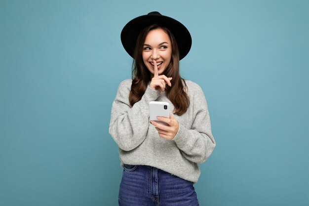 Mulher jovem e atraente sorridente usando chapéu preto e suéter cinza segurando o smartphone, olhando para o lado, mostrando shhh gesto isolado no fundo.