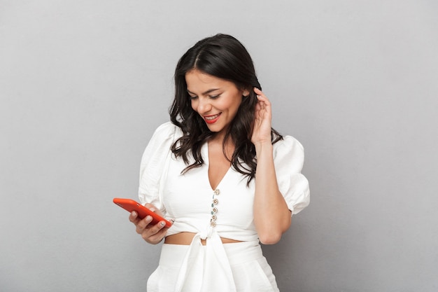 Mulher jovem e atraente sorridente, vestindo uma roupa de verão, em pé, isolada sobre uma parede cinza, usando um telefone celular