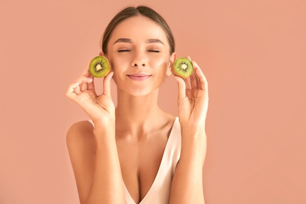 Mulher jovem e atraente sorrindo com os olhos fechados e segurando um kiwi nas mãos sobre um fundo bege