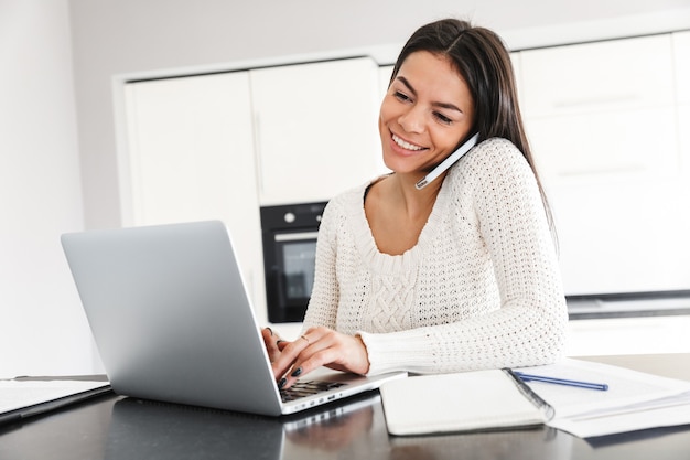 Mulher jovem e atraente trabalhando com um laptop e documentos enquanto está sentado na cozinha, usando um telefone celular