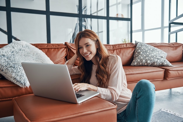 Mulher jovem e atraente usando laptop e sorrindo enquanto passa um tempo em casa