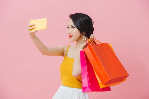Mulher jovem e atraente usando roupa de verão em pé, isolada sobre um fundo rosa, tirando uma selfie, carregando sacolas de compras