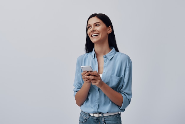 Foto mulher jovem e atraente usando um telefone inteligente e sorrindo em pé contra uma parede cinza