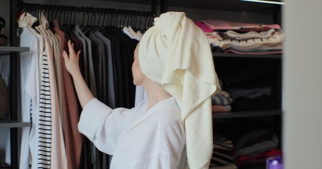 Foto mulher jovem e atraente vestindo um roupão de banho e uma toalha escolhendo suas roupas no armário no quarto em casa