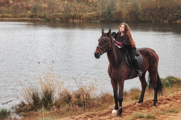 Mulher jovem e bonita a cavalo na floresta de outono pelo lago