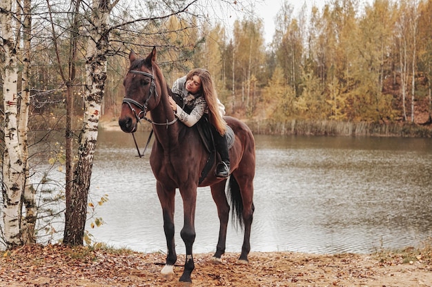 Mulher jovem e bonita a cavalo na floresta de outono pelo lago